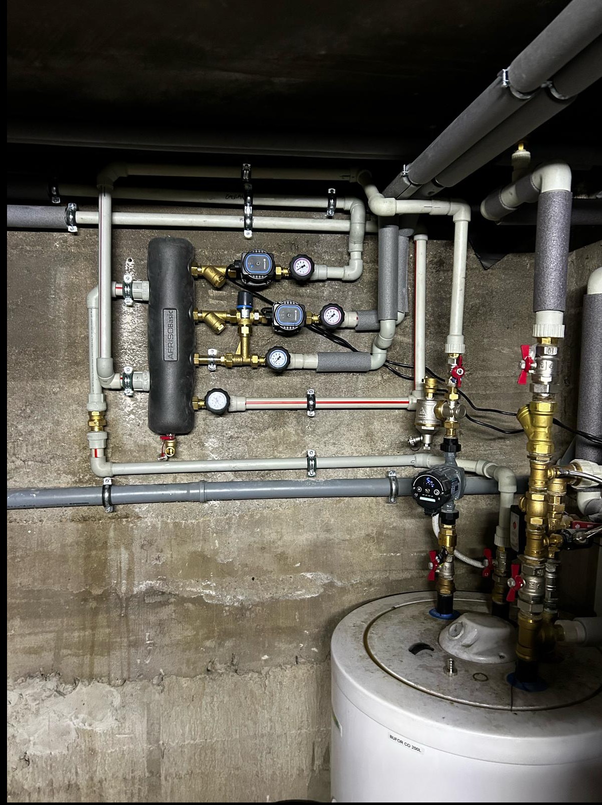 Złożona instalacja hydrauliczna z rurami, zaworami, manometrami i zbiornikiem buforowym, zamontowana na surowej, betonowej ścianie. Widoczne elementy izolacji termicznej.