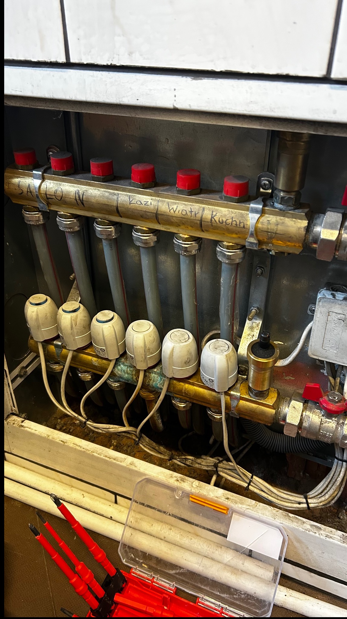 Rozdzielacz ogrzewania podłogowego z rurami i zaworami, opisany nazwami pomieszczeń (Salon, Kazimierz, Wiatr, Kuchnia). Widoczne narzędzia hydrauliczne.