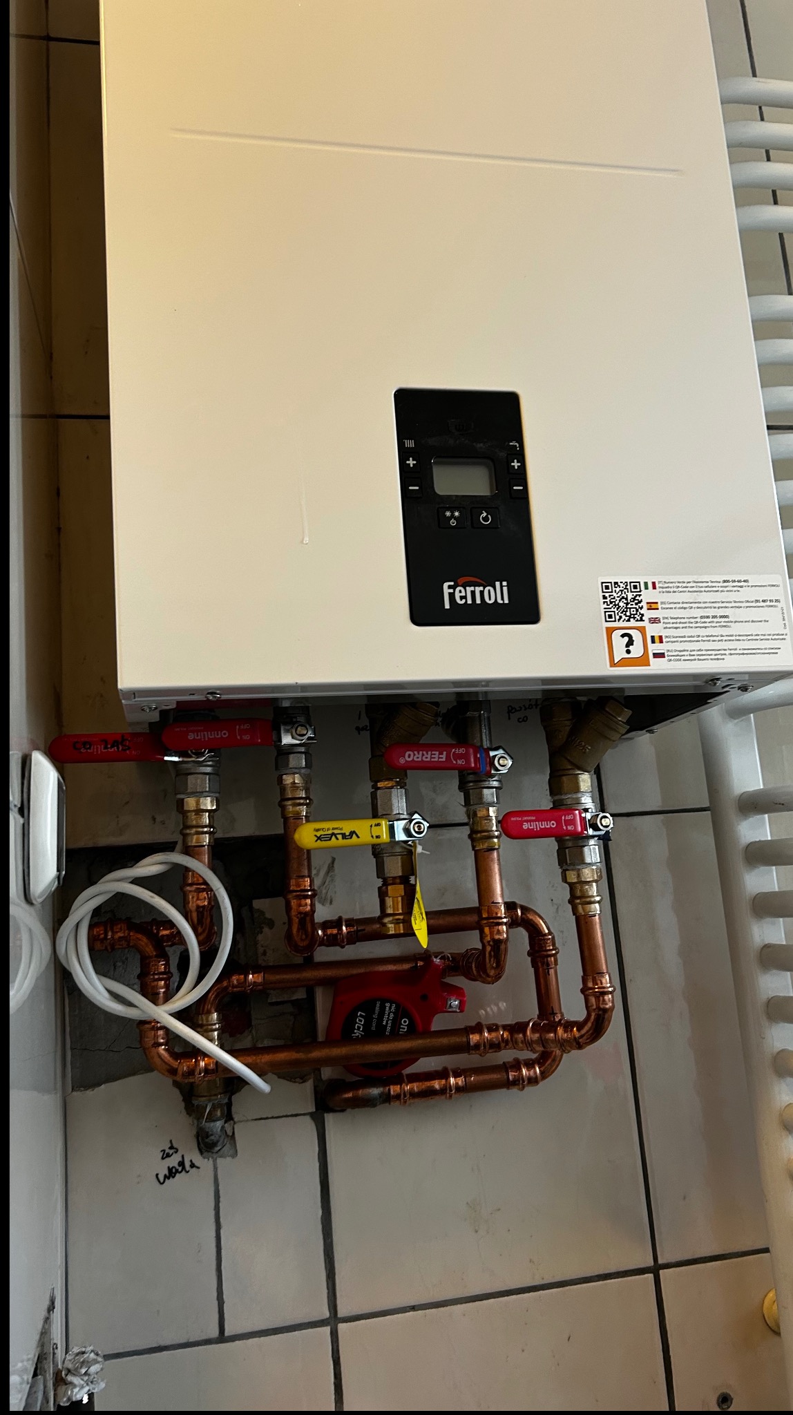 Biały piec gazowy Ferroli z miedzianą instalacją, zaworami (czerwony, żółty) i pompą obiegową. Widoczne płytki ceramiczne na ścianie i fragment grzejnika.