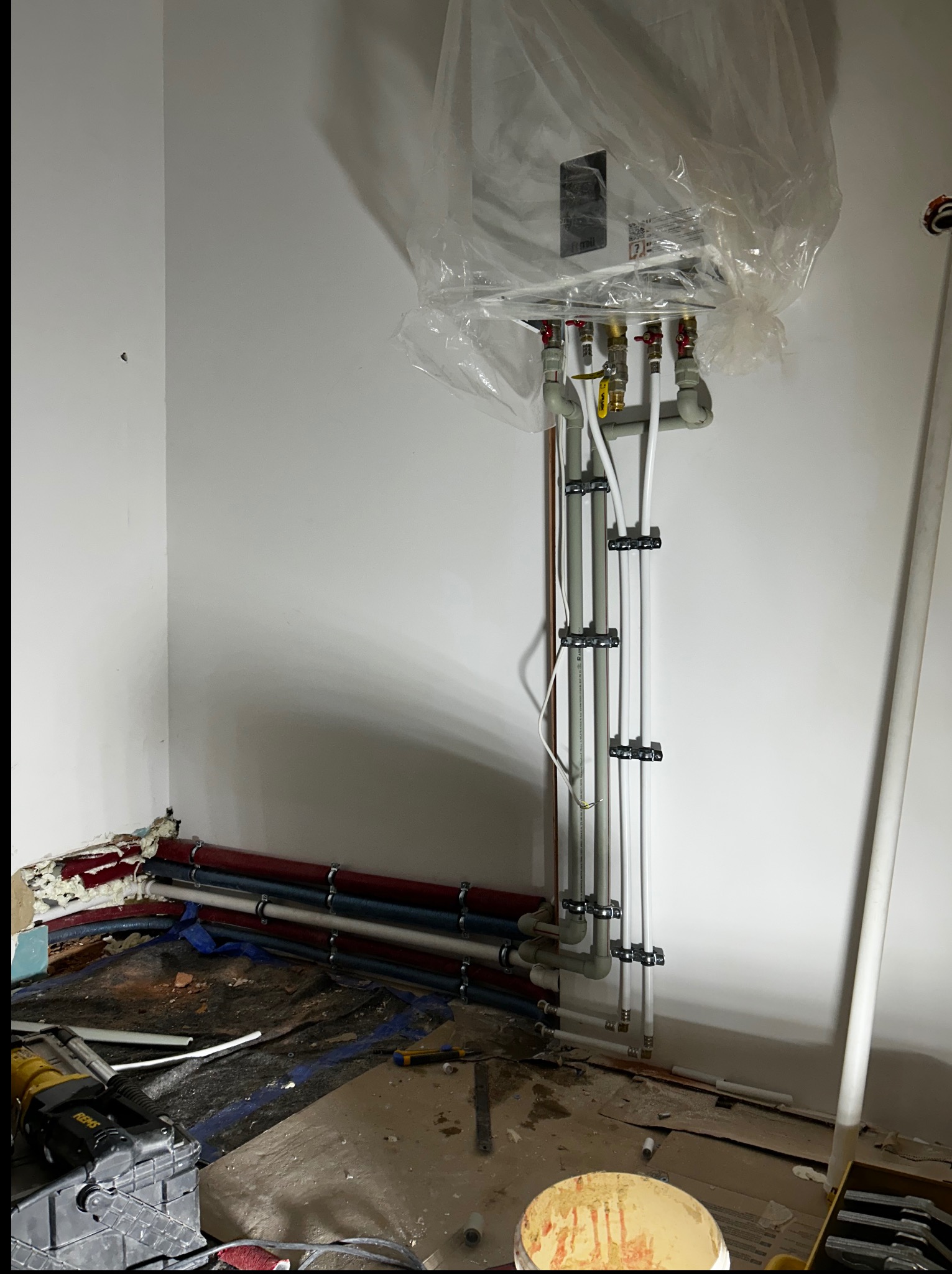 Nowy piec gazowy w folii ochronnej, z podłączonymi rurami instalacji grzewczej. Widoczne narzędzia i fragment podłogi w trakcie montażu.