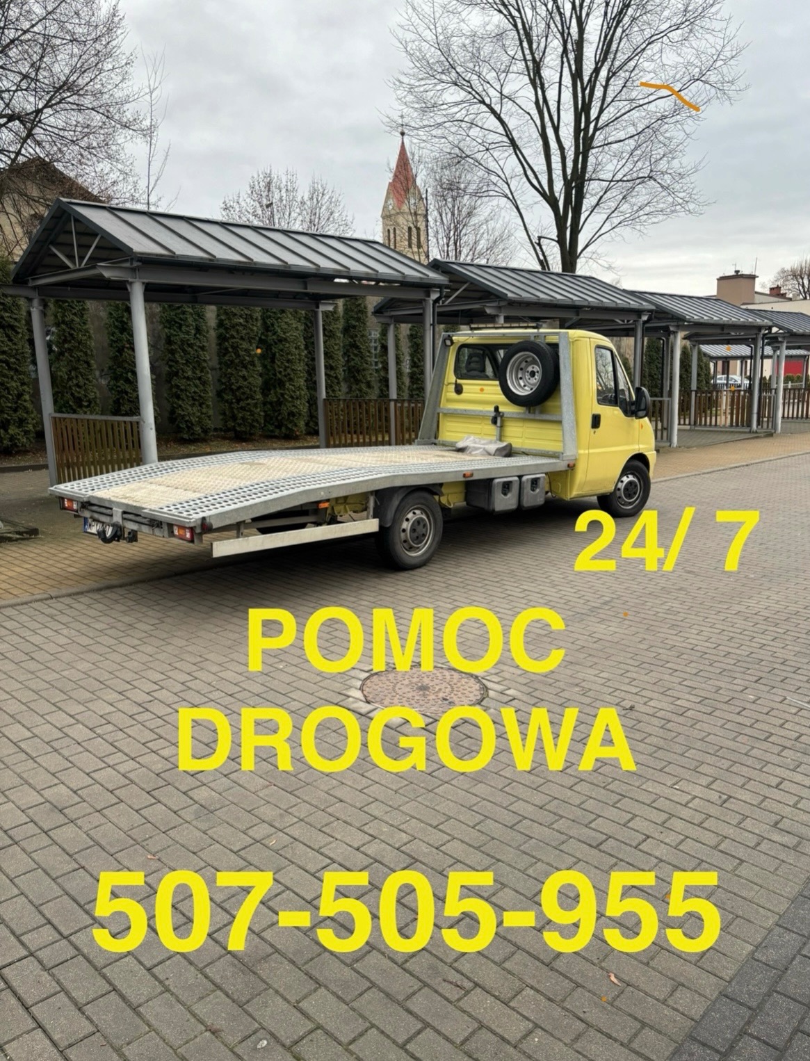 Żółty samochód pomocy drogowej z platformą transportową na kostce brukowej. Widoczny numer telefonu i napis 'Pomoc Drogowa 24/7'. W tle budynki.