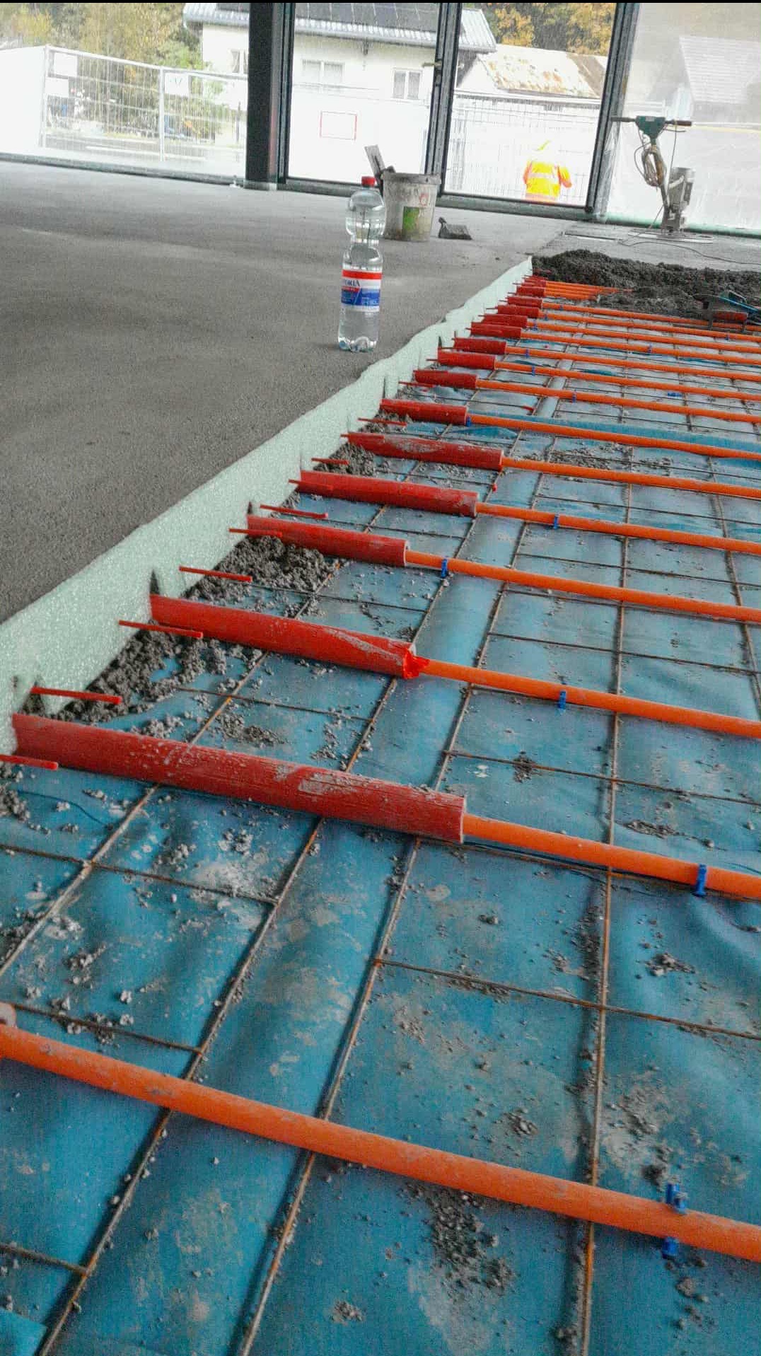 Montaż ogrzewania podłogowego z rurkami w kolorze pomarańczowym, częściowo zalane jastrychem, widoczne mocowania i folia izolacyjna.