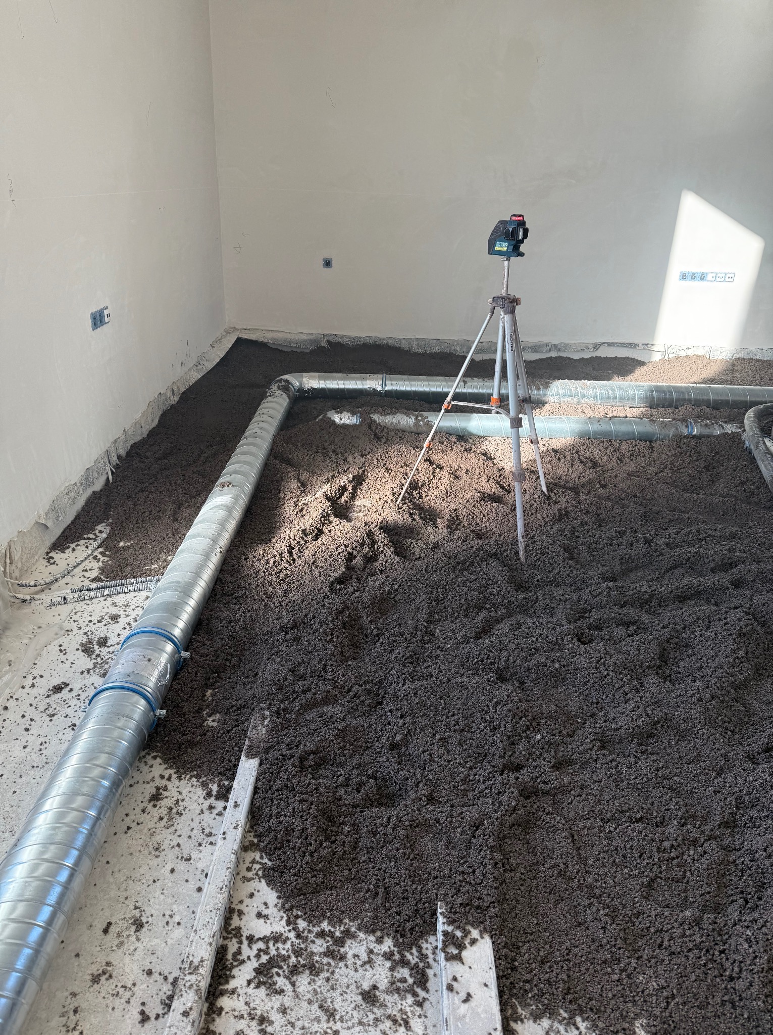Wnętrze z ułożonymi rurami wentylacyjnymi i przygotowaną, ciemną masą na podłodze, gotową do wylania posadzki. Widoczny niwelator laserowy na statywie.