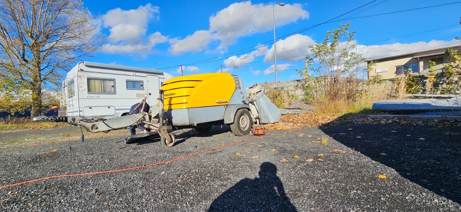 Żółto-szara maszyna do jastrychu Meister na żwirowym podłożu, obok przyczepa kempingowa. W tle budynek i błękitne niebo z chmurami.