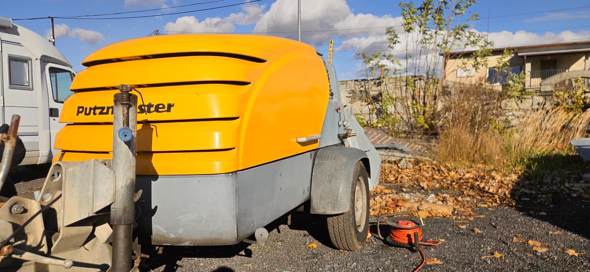 Żółto-szara maszyna Putzmeister na przyczepie, widoczna z boku, z pomarańczowym przedłużaczem na ziemi. W tle dom i roślinność.