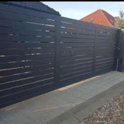 Nowoczesna, przesuwna brama wjazdowa z poziomymi lamelami w antracytowym kolorze, prowadząca do domu z czerwoną dachówką, widoczna z poziomu chodnika.