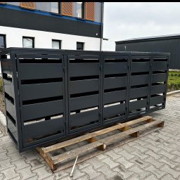 HUBWELD SPÓŁKA Z OGRANICZONĄ ODPOWIEDZIALNOŚCIĄ - Metalowa obudowa śmietnikowa z poziomymi szczelinami, pomalowana na szaro, stoi na palecie na tle budynku z szarą elewacją i kostki brukowej. Minimalistyczny design.