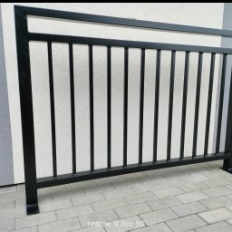 HUBWELD SPÓŁKA Z OGRANICZONĄ ODPOWIEDZIALNOŚCIĄ - Czarna, metalowa balustrada zewnętrzna z pionowymi prętami na tle szarej ściany i kostki brukowej. Widoczne detale spawów i mocowania do podłoża.