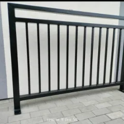 Czarna, metalowa balustrada zewnętrzna z pionowymi prętami na tle szarej ściany i kostki brukowej. Widoczne detale spawów i mocowania do podłoża.