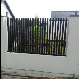 HUBWELD SPÓŁKA Z OGRANICZONĄ ODPOWIEDZIALNOŚCIĄ - Nowoczesne ogrodzenie z czarnych, pionowych metalowych elementów osadzonych w białym, murowanym cokole. W tle budynek z szarym dachem i zielona roślinność.