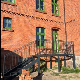 HUBWELD SPÓŁKA Z OGRANICZONĄ ODPOWIEDZIALNOŚCIĄ - Stalowe schody z balustradą o geometrycznym wzorze, prowadzące do wejścia w ceglanym budynku z zielonymi ramami okiennymi. U dołu sterta cegieł.