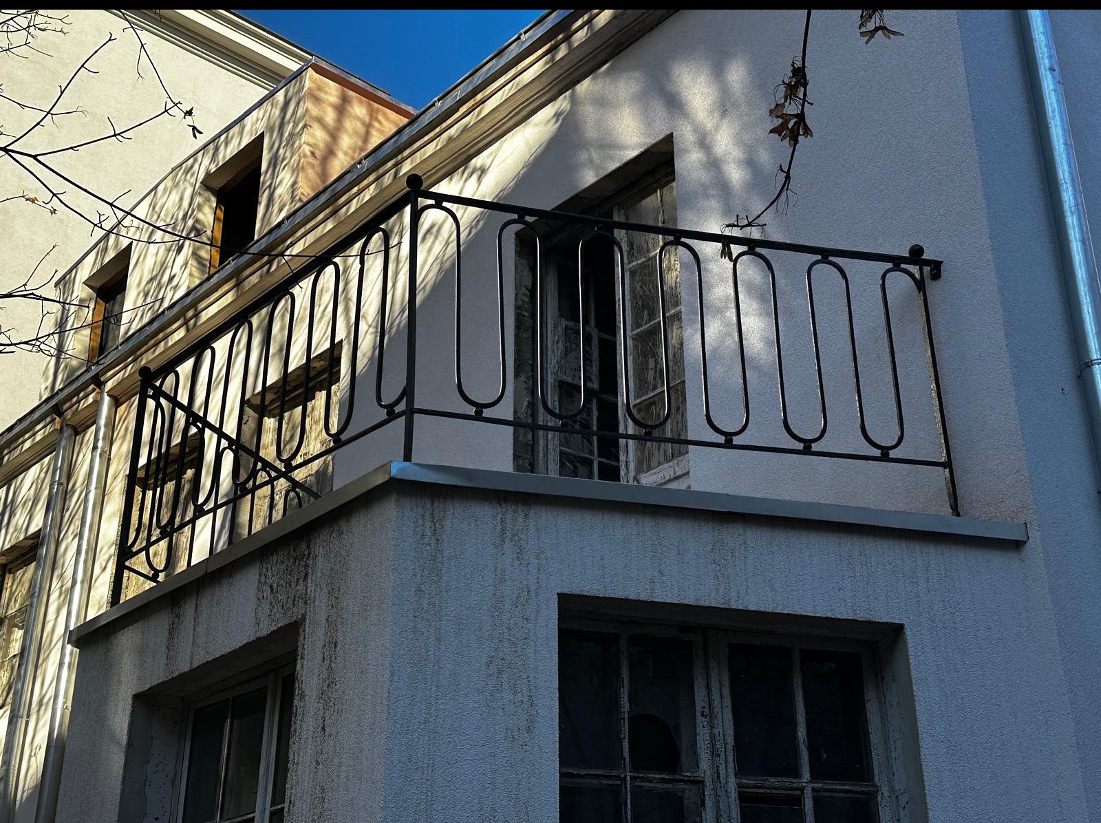 Czarna, metalowa balustrada balkonowa z owalnymi prętami na tle jasnej elewacji budynku z widocznymi oknami i fragmentami drzew. Ujęcie z dołu, w słoneczny dzień.