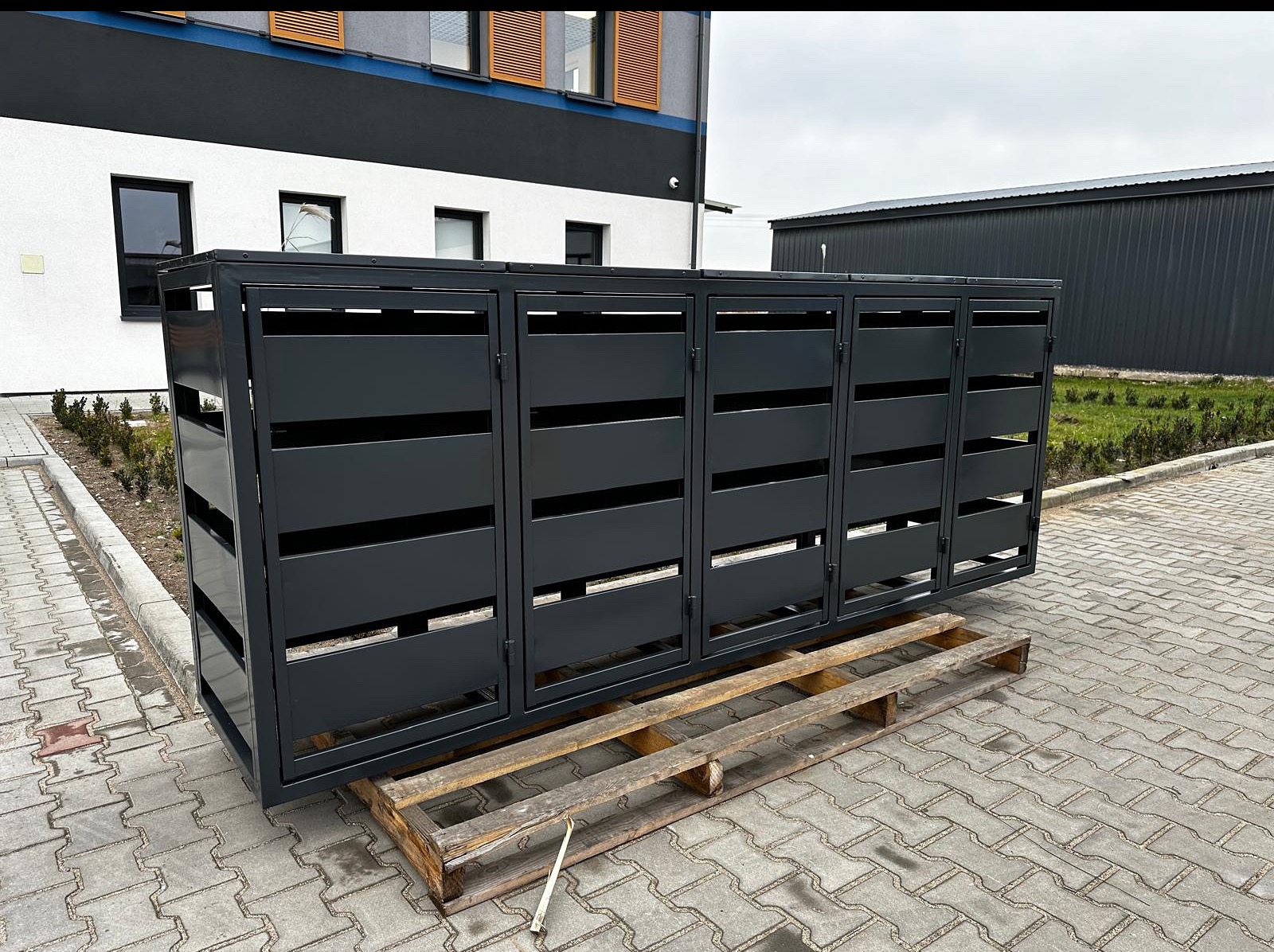 Metalowa obudowa śmietnikowa z poziomymi szczelinami, pomalowana na szaro, stoi na palecie na tle budynku z szarą elewacją i kostki brukowej. Minimalistyczny design.