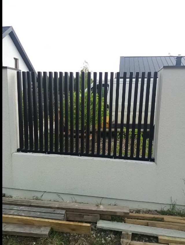 Nowoczesne ogrodzenie z czarnych, pionowych metalowych elementów osadzonych w białym, murowanym cokole. W tle budynek z szarym dachem i zielona roślinność.