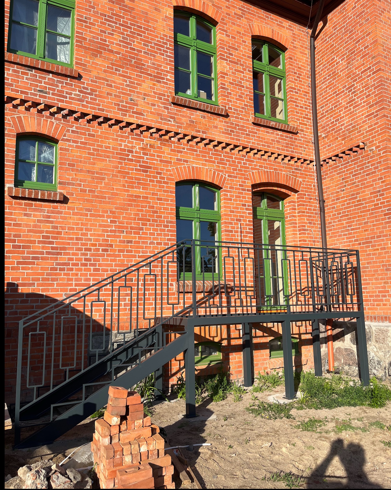 Stalowe schody z balustradą o geometrycznym wzorze, prowadzące do wejścia w ceglanym budynku z zielonymi ramami okiennymi. U dołu sterta cegieł.