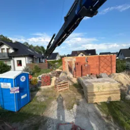 Budowa domu jednorodzinnego: wznoszenie murów z czerwonej cegły. Na pierwszym planie materiały budowlane i przenośna toaleta. W tle domy jednorodzinne.