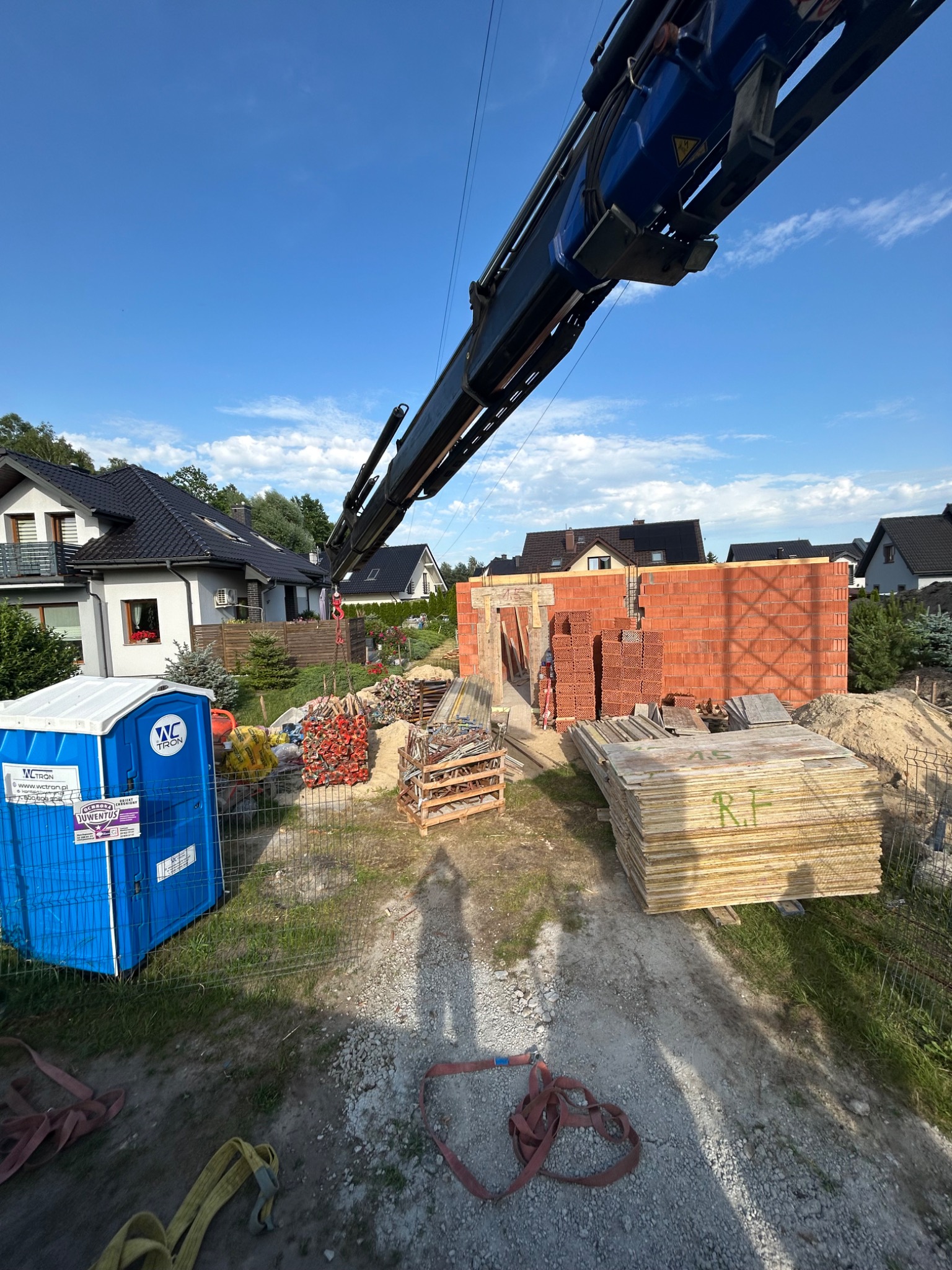 Budowa domu jednorodzinnego: wznoszenie murów z czerwonej cegły. Na pierwszym planie materiały budowlane i przenośna toaleta. W tle domy jednorodzinne.