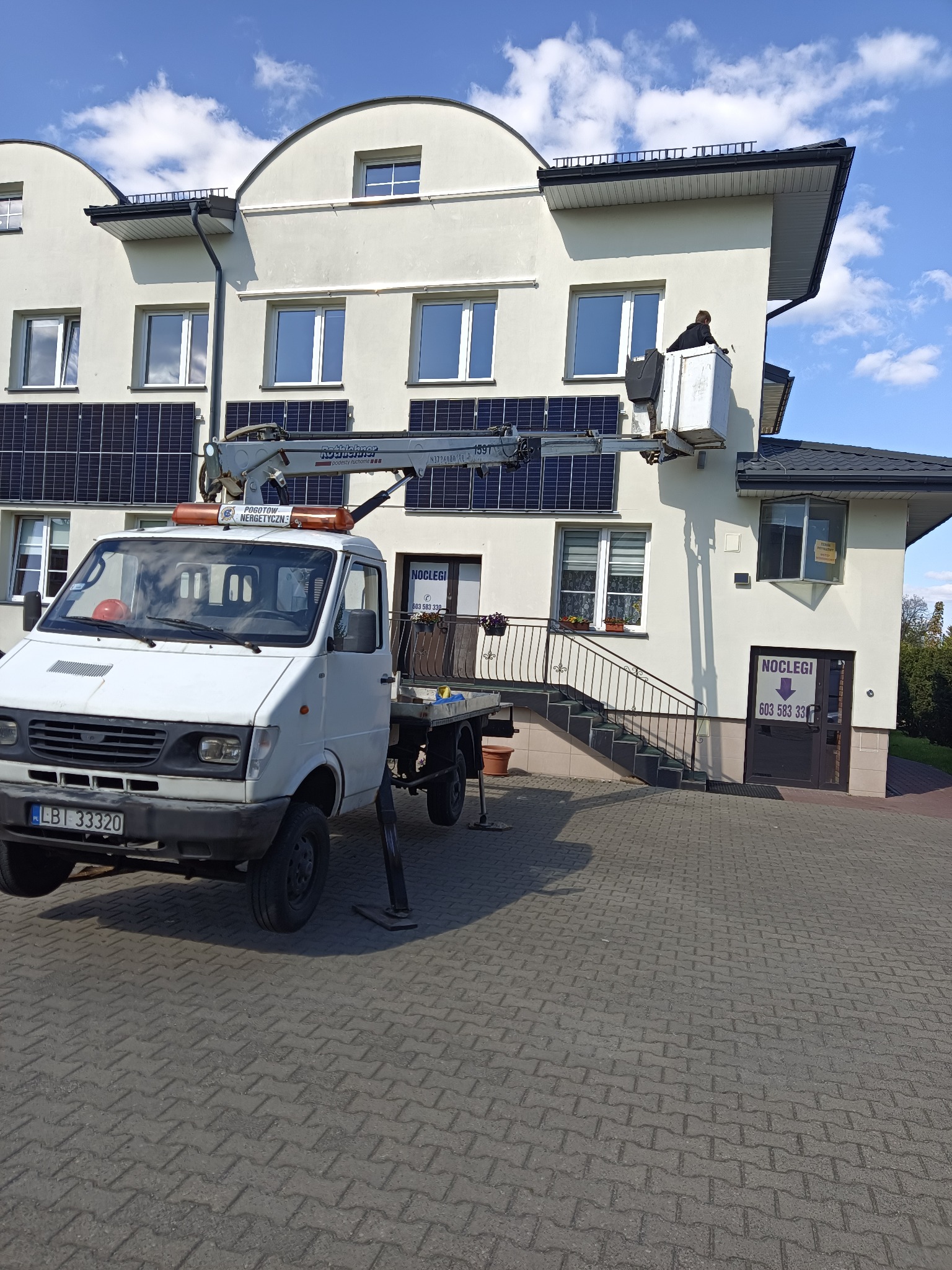 Pracownik w koszu podnośnika na tle budynku z panelami słonecznymi. Na pierwszym planie samochód z napisem 'Pogotowie Energetyczne'.