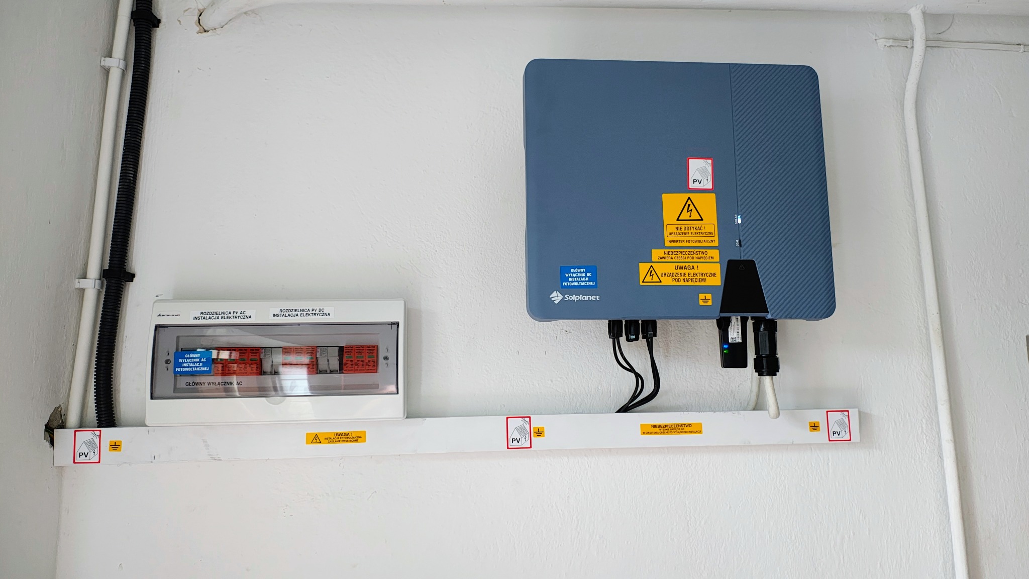 Szary inwerter fotowoltaiczny Solplanet zamontowany na białej ścianie obok rozdzielnicy elektrycznej. Widoczne przewody i oznaczenia ostrzegawcze.
