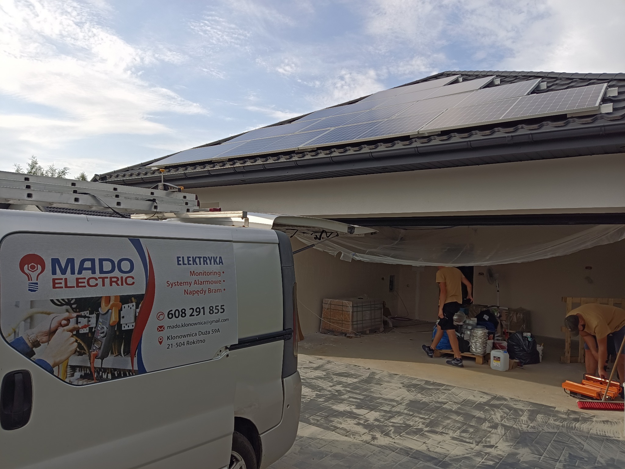 Montaż paneli słonecznych na dachu domu, widoczny samochód firmy Mado Electric z reklamą usług elektrycznych, alarmów i automatyki bram. Ekipa montuje kostkę brukową.