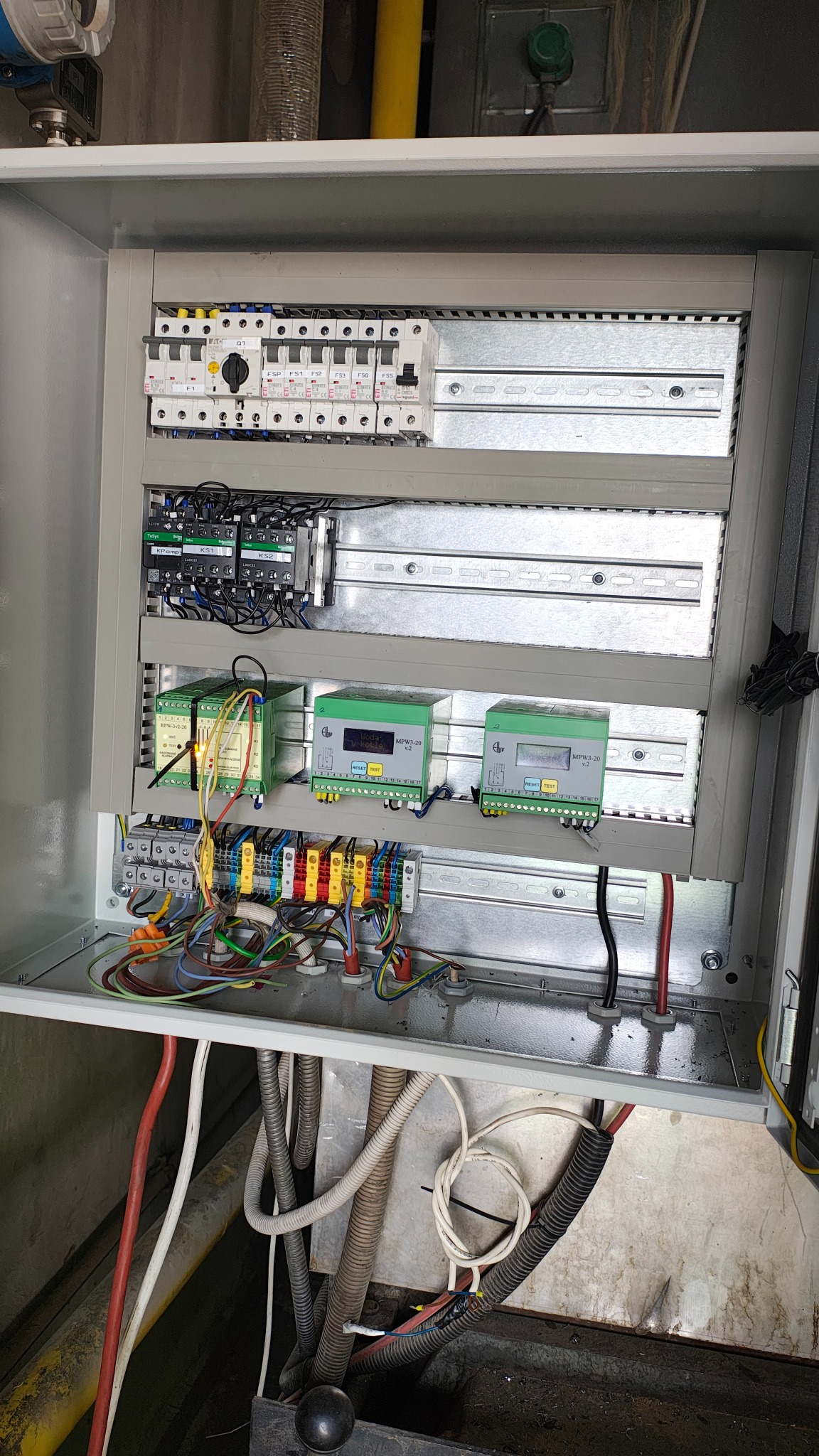 Otwarta szafa sterownicza z widocznymi bezpiecznikami, modułami sterującymi i okablowaniem. Kolorowe przewody podłączone do listew zaciskowych. Widoczne elementy instalacji.