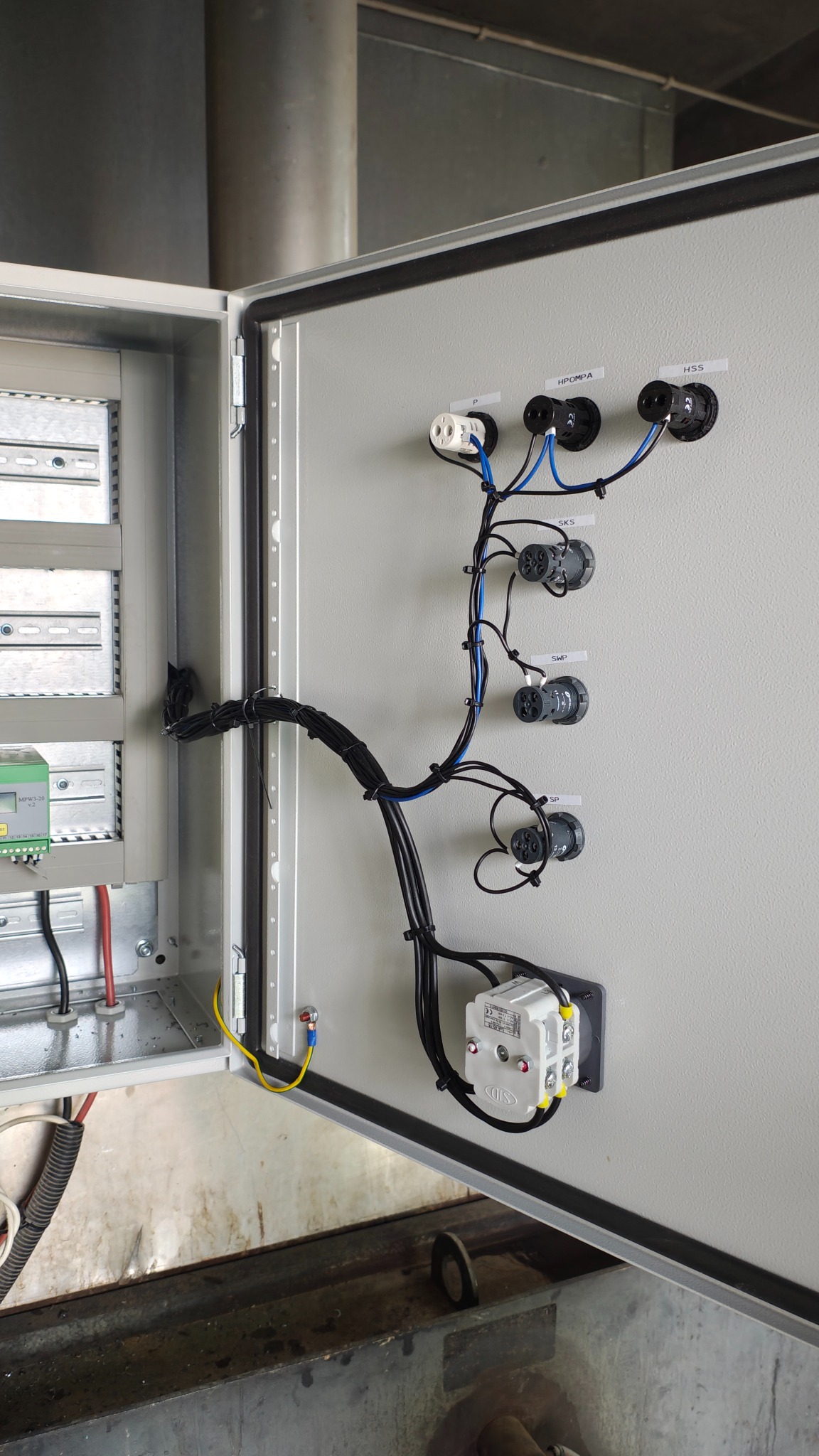 Otwarta szafa sterownicza z widocznymi elementami instalacji elektrycznej, w tym przewodami i gniazdami oznaczonymi etykietami. Widoczne okablowanie i moduły sterujące.