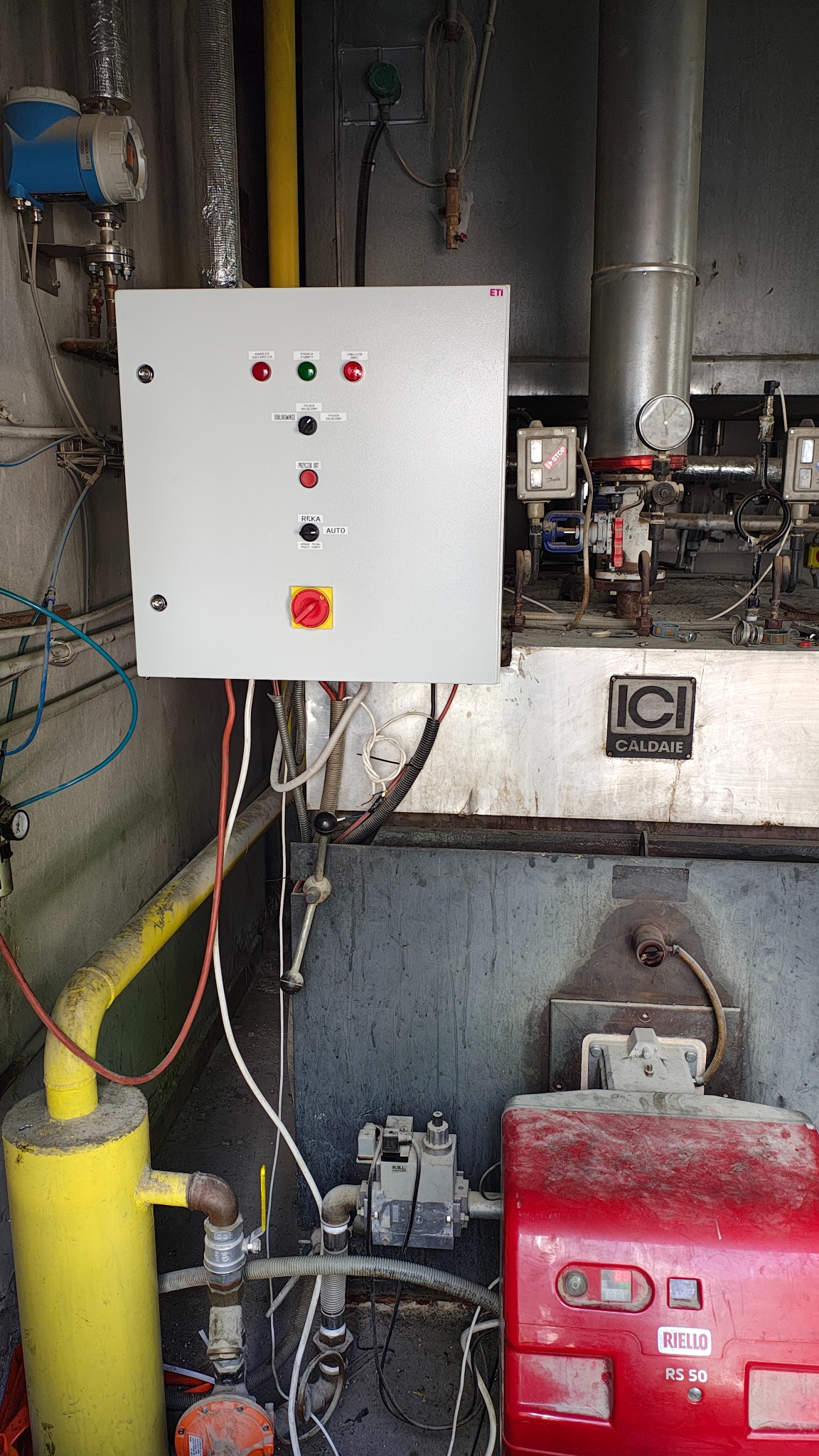 Panel sterowania z przyciskami i przełącznikami, zamontowany obok kotła przemysłowego z widocznymi rurami i przewodami. Elementy instalacji w kotłowni.