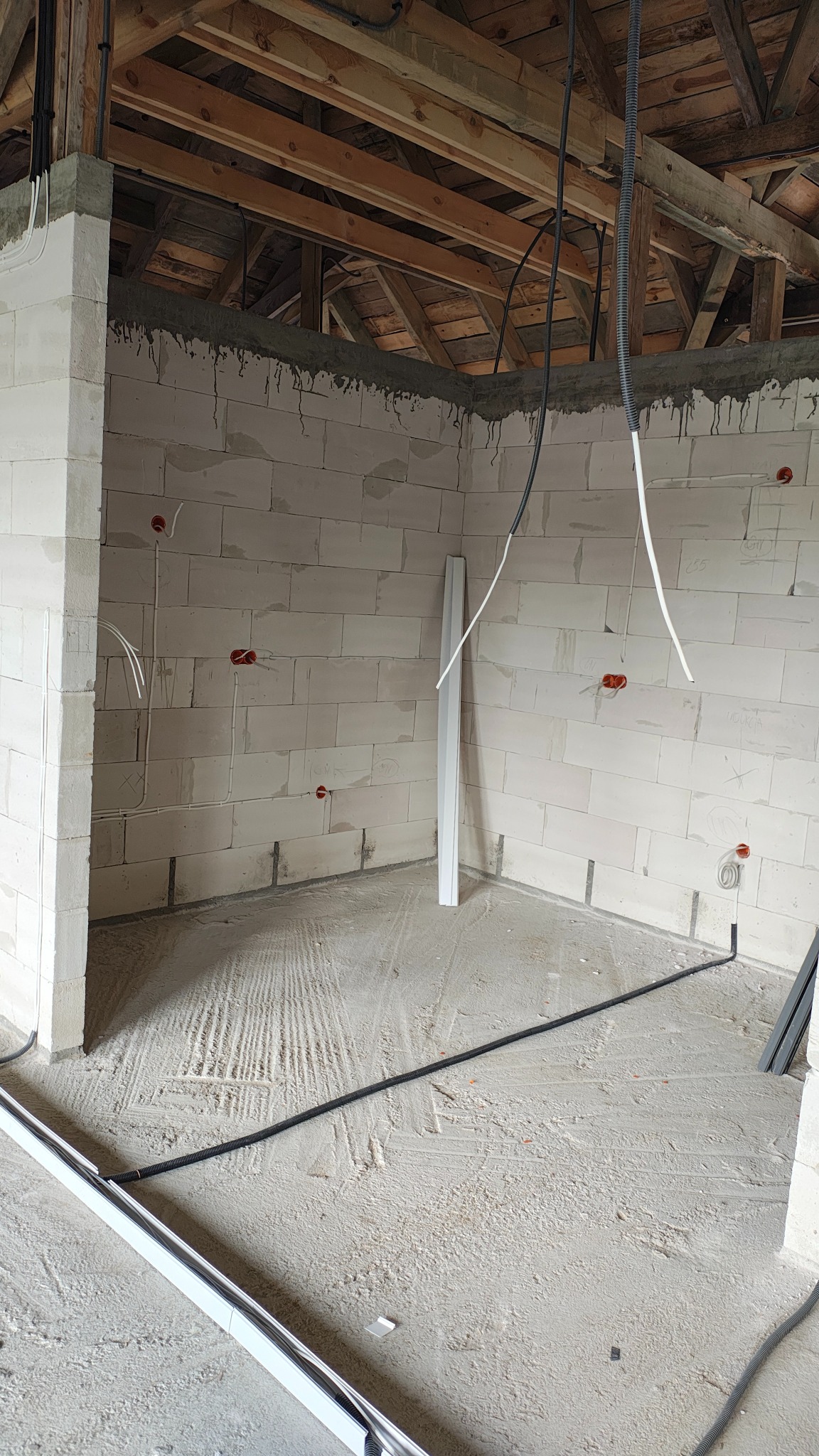 Wnętrze w surowym stanie z instalacją elektryczną w ścianach z betonu komórkowego. Widoczne kable i puszki elektryczne, oraz fragmenty rur instalacyjnych na podłodze.