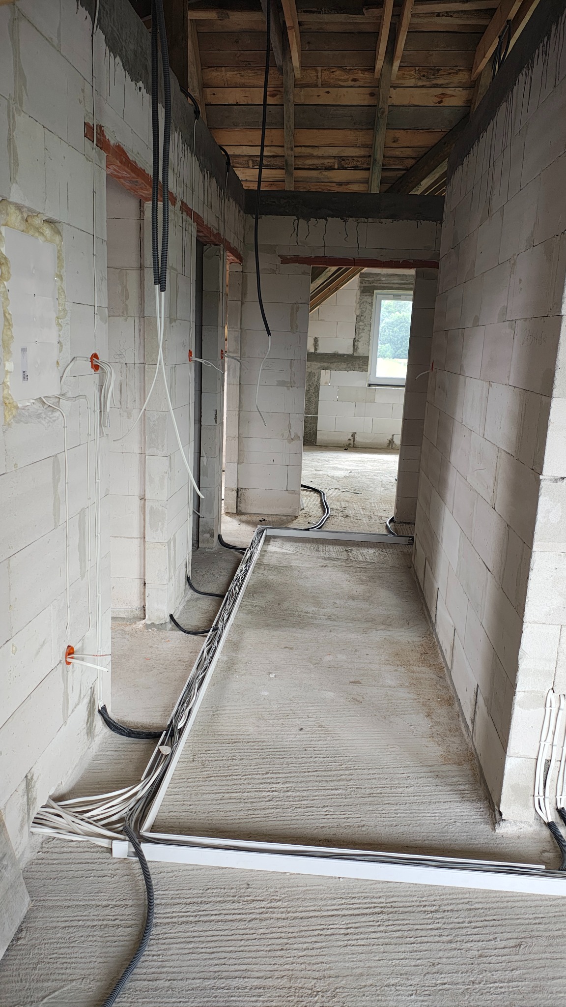 Wnętrze w trakcie budowy z widocznymi przewodami elektrycznymi ułożonymi w korytkach podłogowych i wyprowadzonymi ze ścian. Surowy stan, ściany z białego betonu komórkowego.