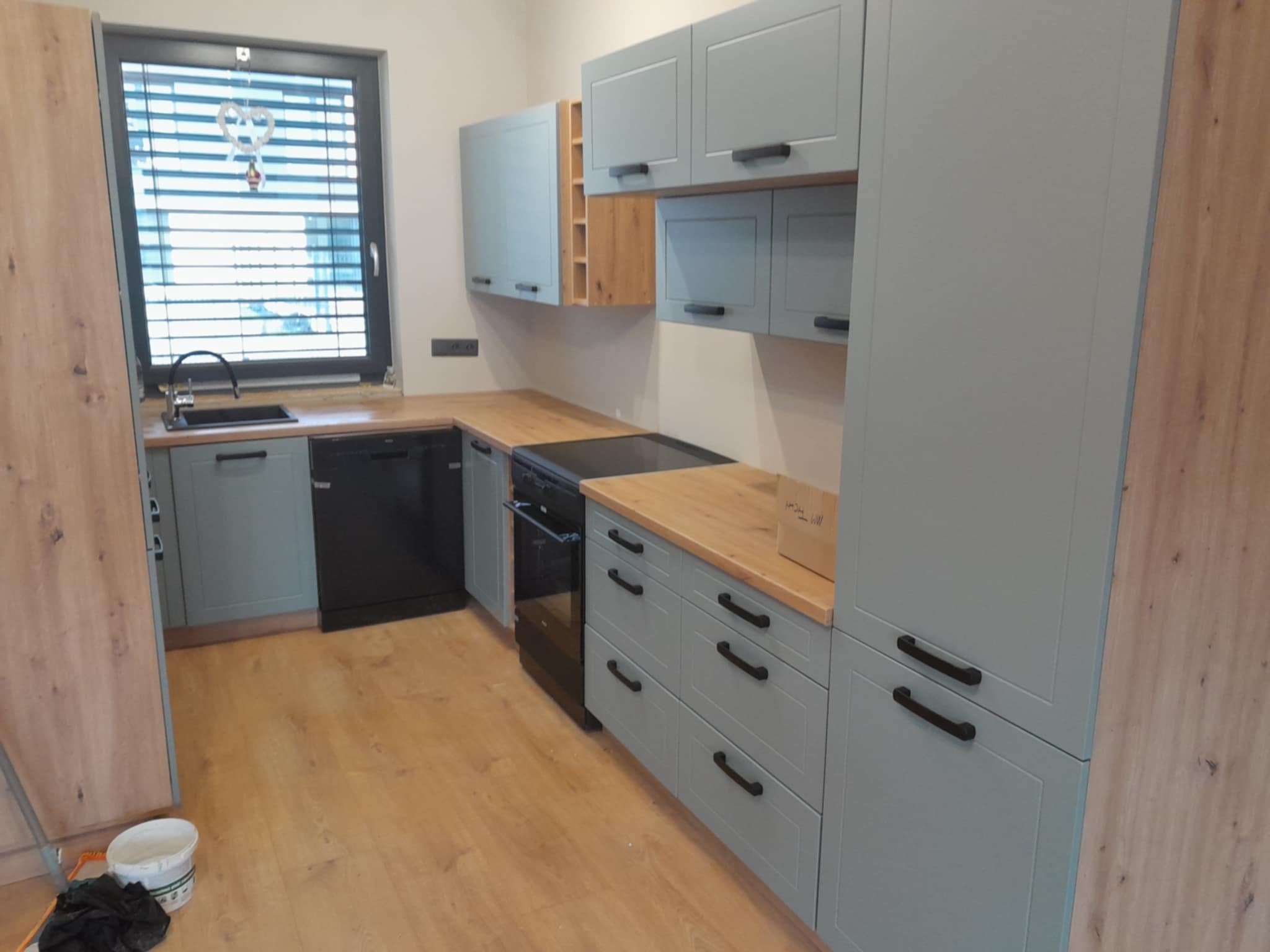 Kompletna kuchnia w odcieniach szarości z drewnianymi blatami i akcentami, widok na okno z żaluzjami i podłogę z paneli, kubełek z farbą w rogu.