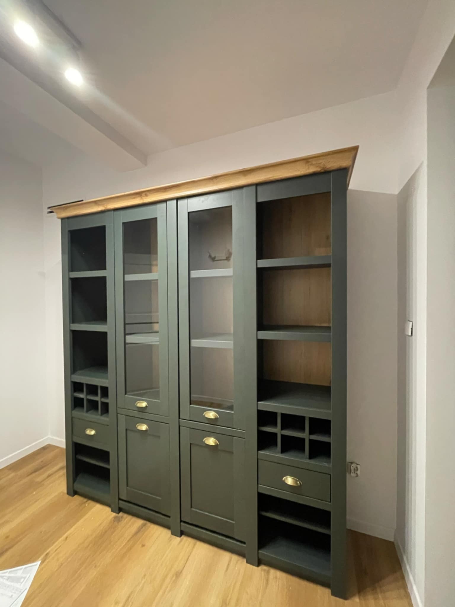 Zabudowa meblowa w kolorze oliwkowym z drewnianym blatem, z przeszklonymi frontami, widoczne półki i szuflady. Ustawiona na parkiecie, w tle biała ściana.