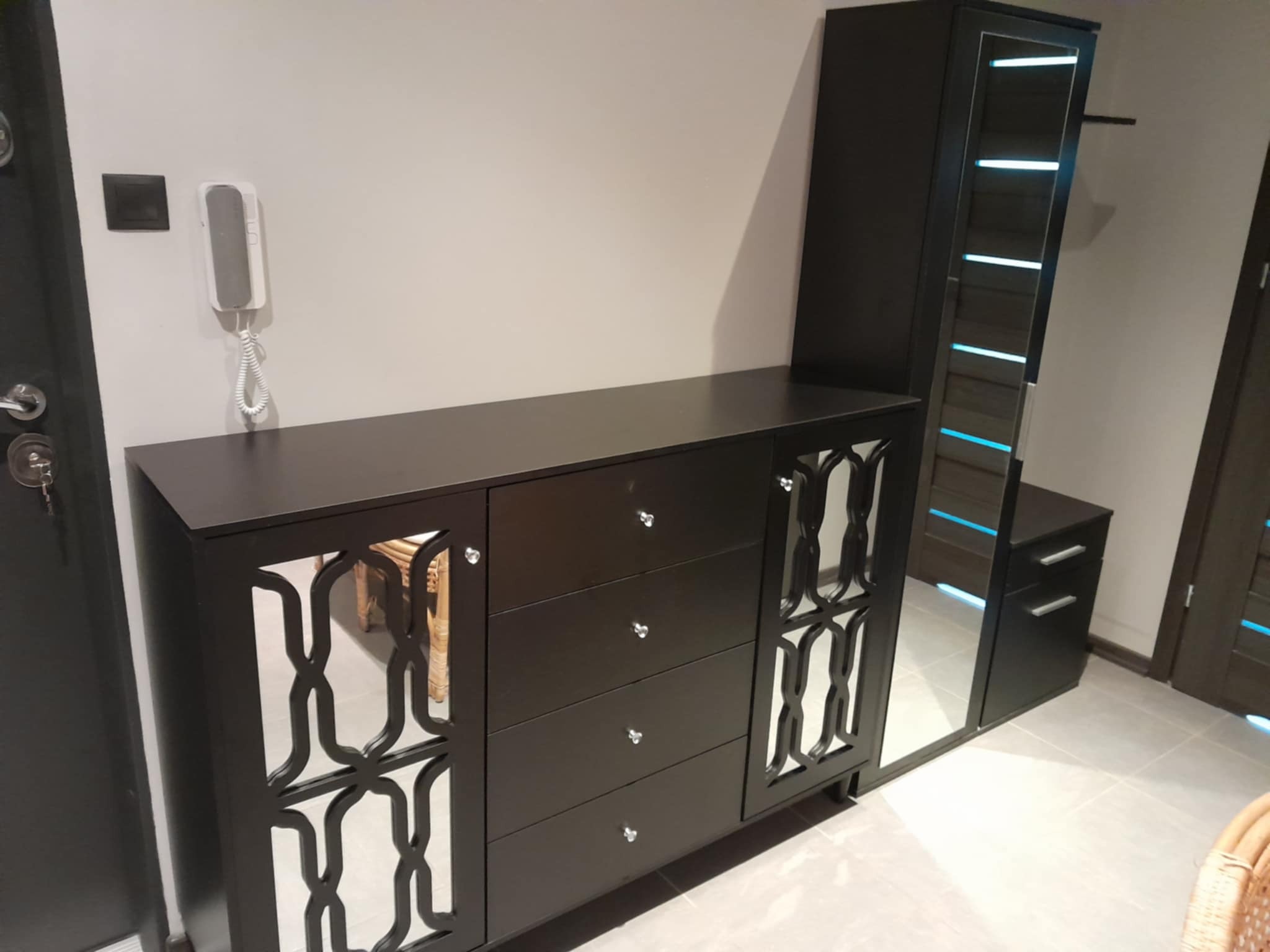 Ciemny komplet mebli w przedpokoju z lustrami i wzorami. Widoczny fragment drzwi wejściowych i domofon. Na podłodze jasne płytki.