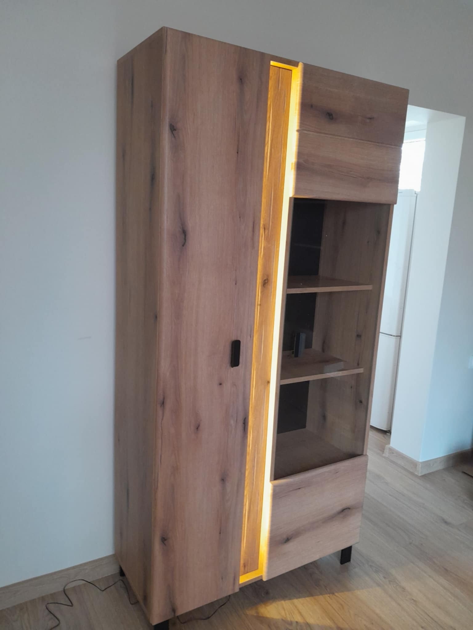 Stojąca szafka z oświetleniem LED, drewnopodobna okleina, widok z boku, na podłodze widoczny kabel zasilający, w tle ściana i fragment lodówki.