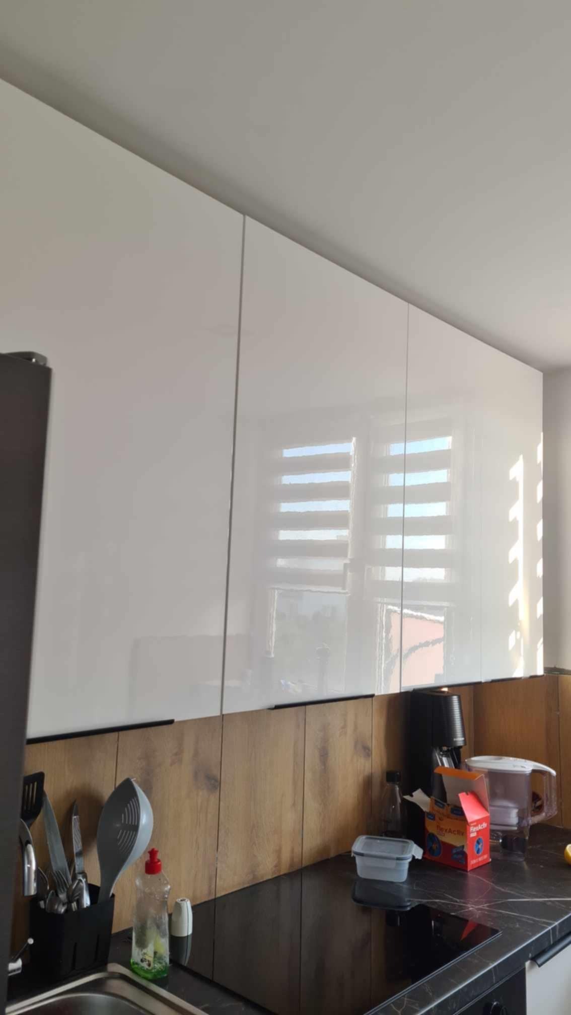 Białe, lakierowane szafki kuchenne pod sufitem, z widocznym fragmentem okna z żaluzjami. Na blacie akcesoria kuchenne i dekoracyjne panele ścienne z efektem drewna.