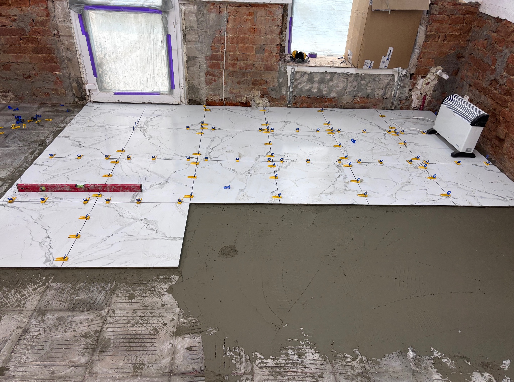 Układanie białych płytek imitujących marmur, z użyciem poziomicy i klipsów poziomujących, na podłodze z częściowo nałożoną warstwą kleju. Widoczne surowe ściany z cegły.