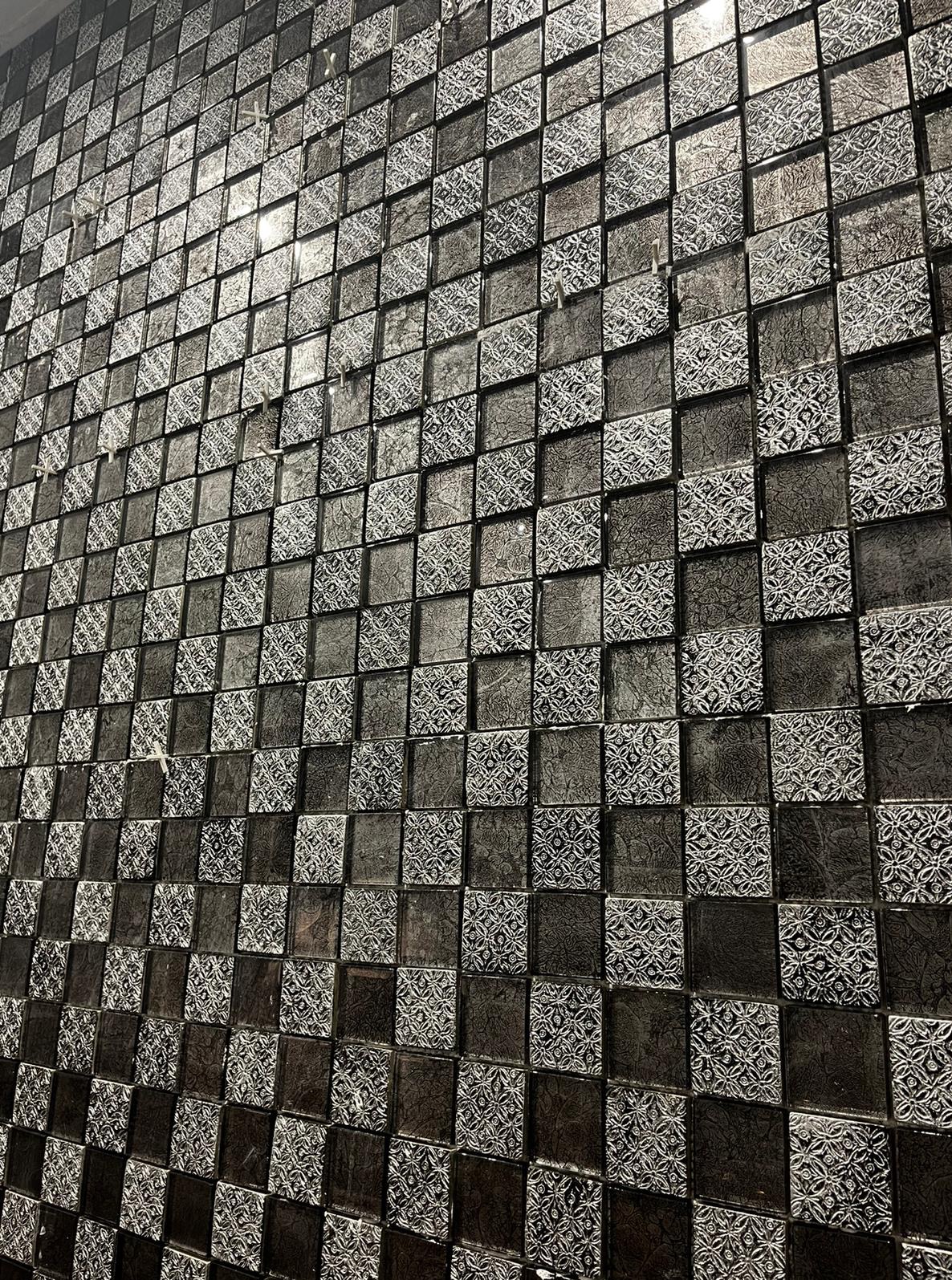 Ściana wykończona mozaiką z kwadratowych płytek w odcieniach szarości i czerni, niektóre z delikatnym wzorem, ułożonych pod kątem, z widocznymi refleksami światła.
