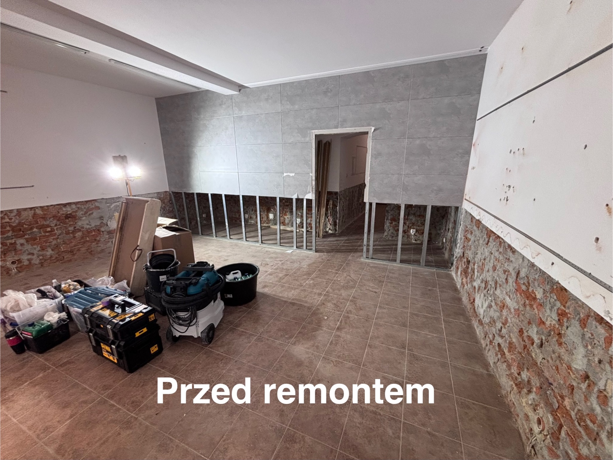 Wnętrze w trakcie remontu: częściowo położone szare płytki na ścianie, odsłonięta cegła, narzędzia i sprzęt budowlany. Widoczny etap prac przed wykończeniem.