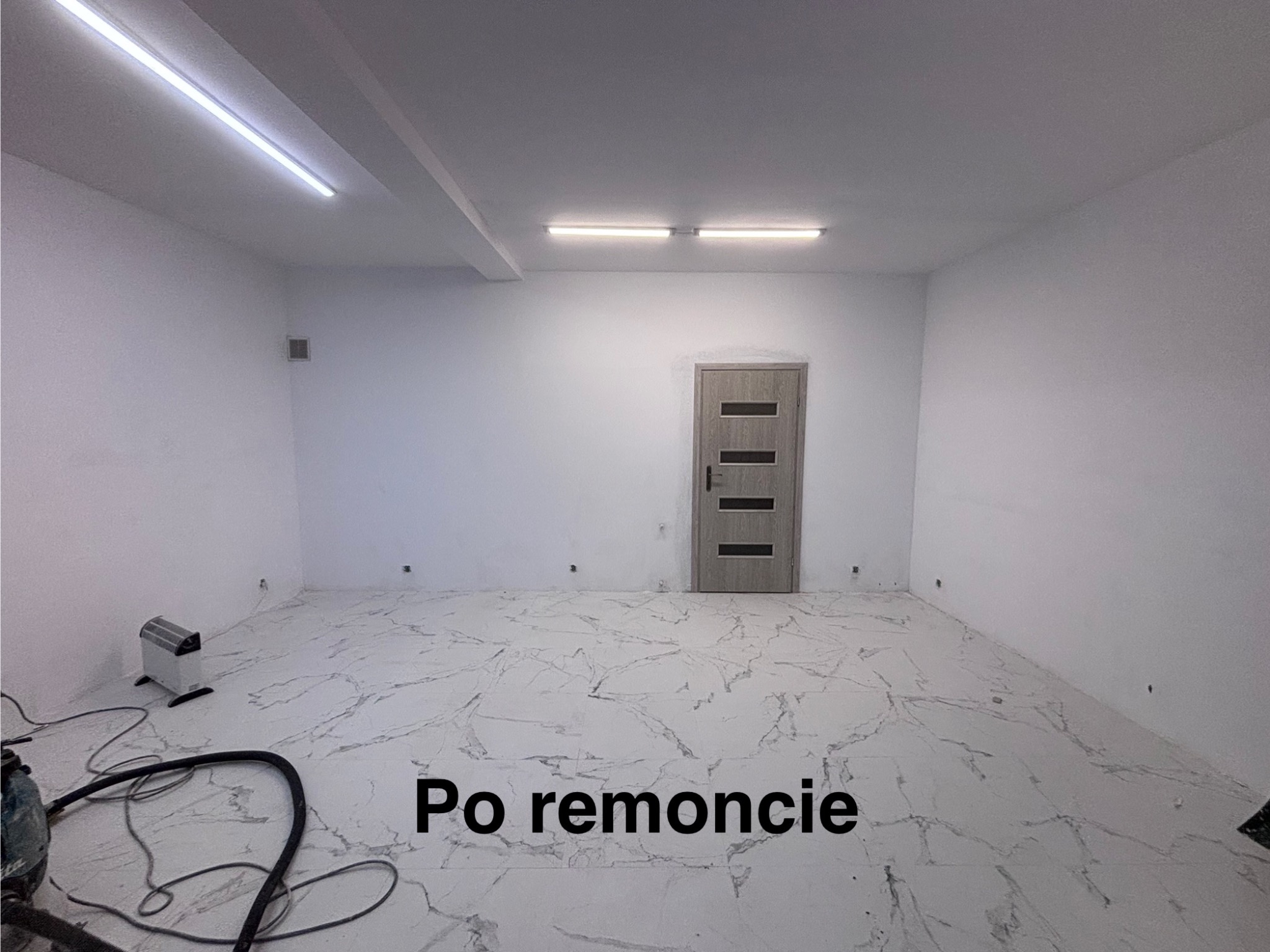 Jasne wnętrze po remoncie: białe ściany, marmurowe płytki podłogowe, szare drzwi z poziomymi przeszkleniami. Widoczne lampy sufitowe i grzejnik elektryczny.