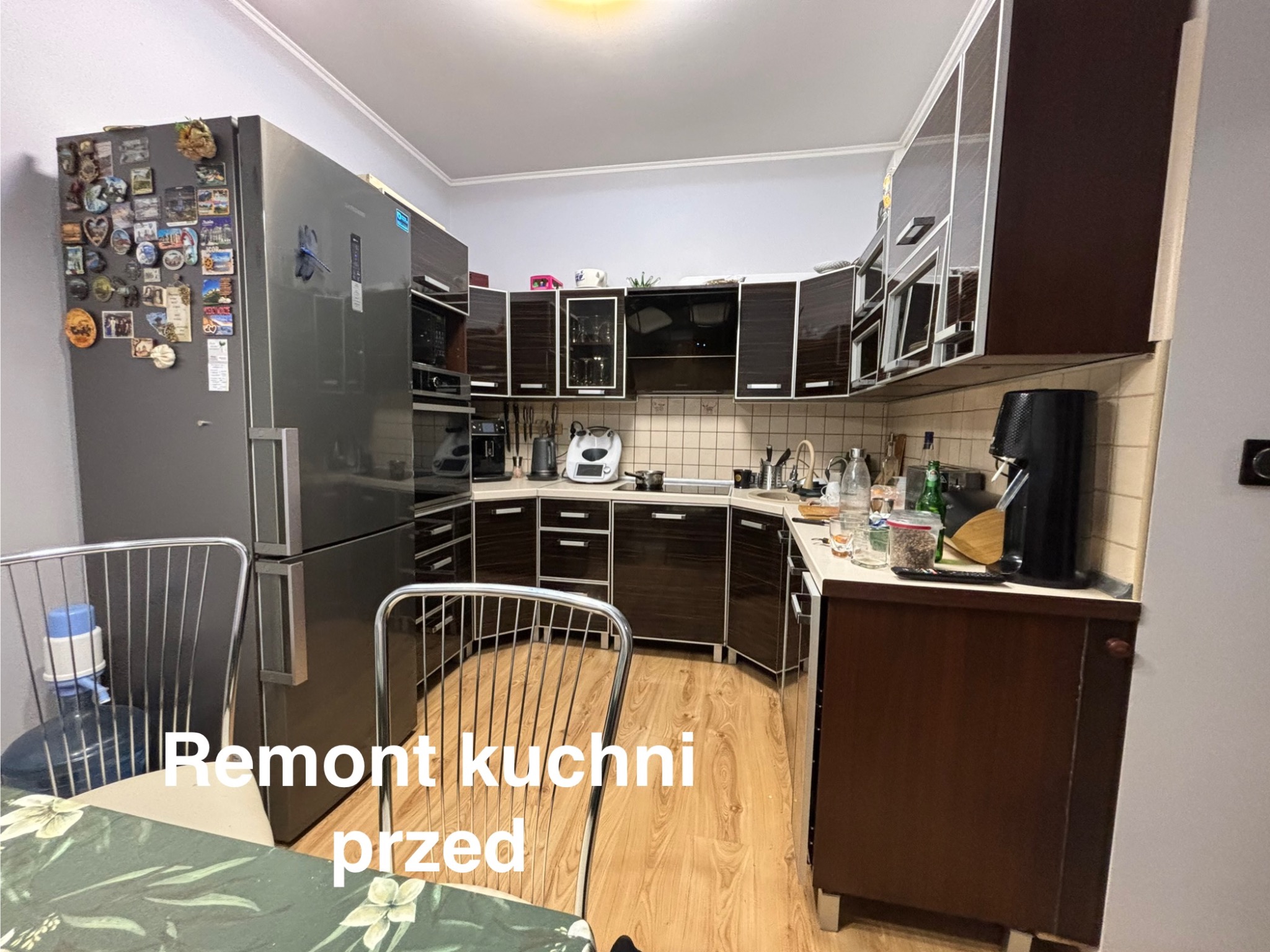 Kuchnia z ciemnymi meblami przed remontem. Widoczny duży, srebrny lodówka z magnesami, drewniana podłoga i krzesła. Ujęcie z perspektywy stołu.