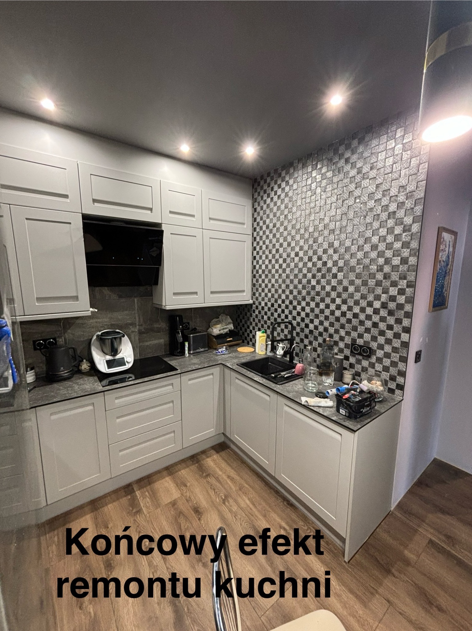 Szara kuchnia po remoncie z mozaiką na ścianie, drewnopodobną podłogą i nowoczesnym sprzętem AGD. Widoczne szafki górne i dolne, okap oraz drobne akcesoria kuchenne.