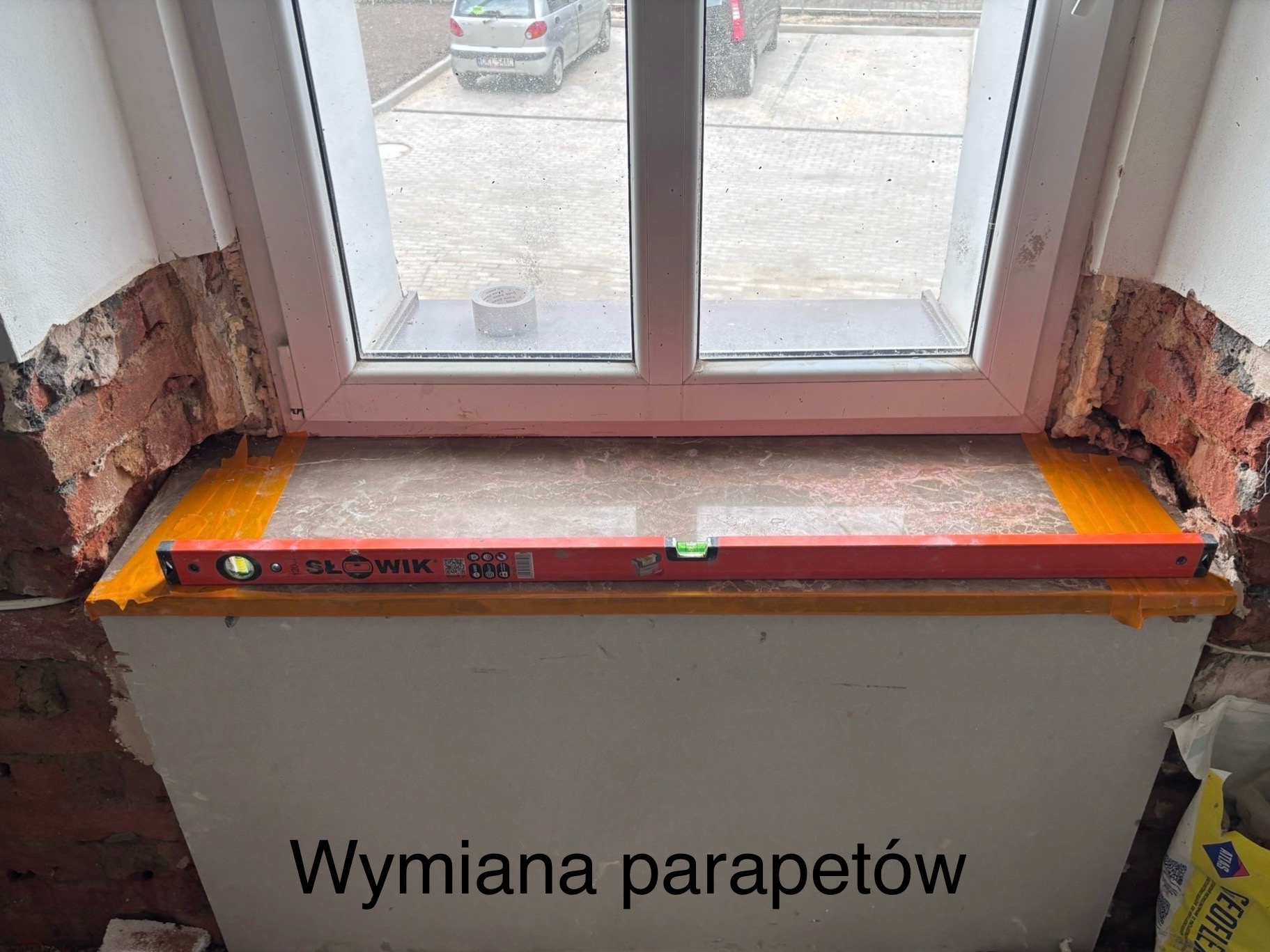 Montaż parapetu okiennego z użyciem poziomicy, widoczne uszkodzenia muru i taśma zabezpieczająca. W tle okno z widokiem na zewnątrz.