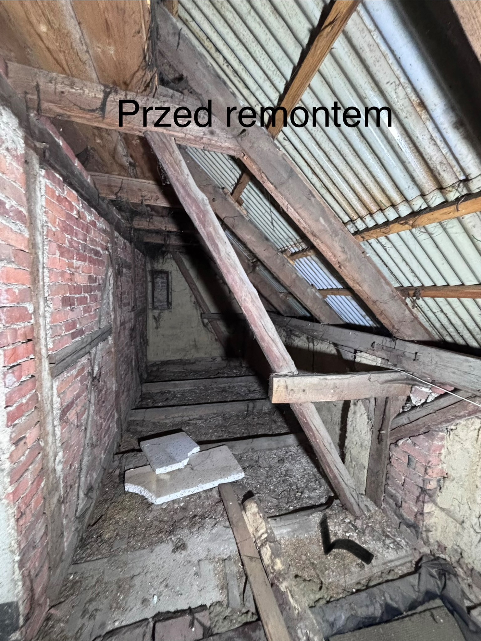 Stare poddasze z odsłoniętą konstrukcją drewnianą i fragmentami ceglanej ściany, widoczne elementy izolacji i gruzu. Zdjęcie 'przed remontem'.