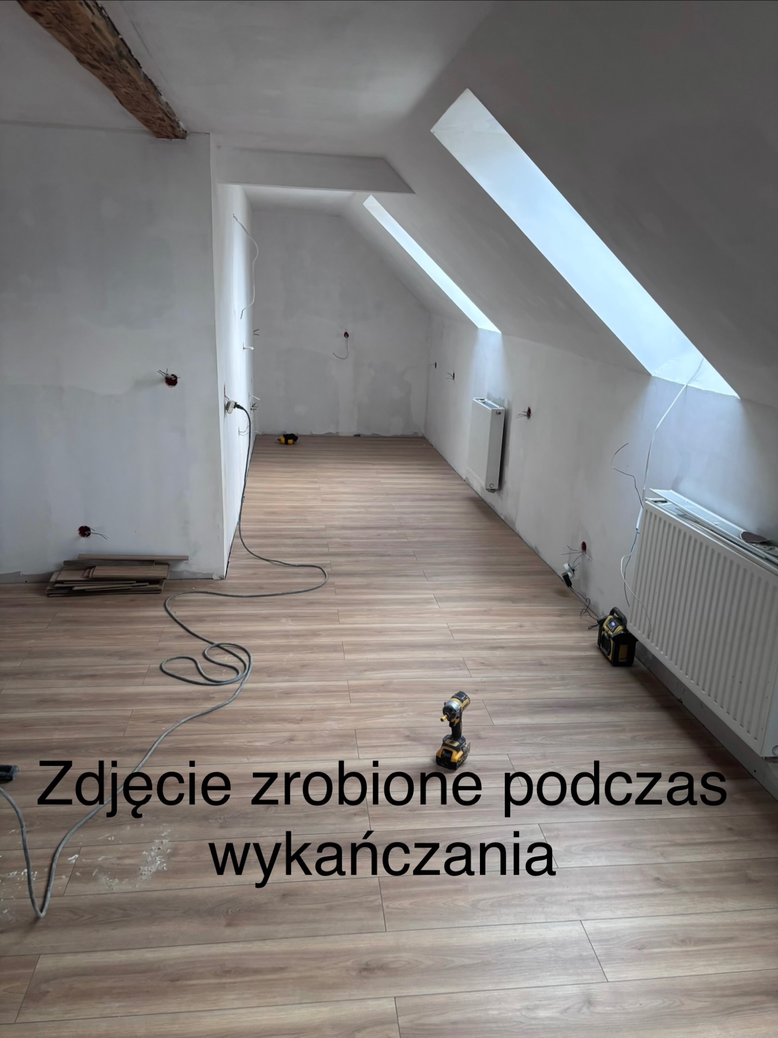 Poddasze z oknami dachowymi w trakcie wykańczania: ułożona podłoga z paneli, grzejniki, ściany przygotowane do malowania, widoczne kable elektryczne i narzędzia.