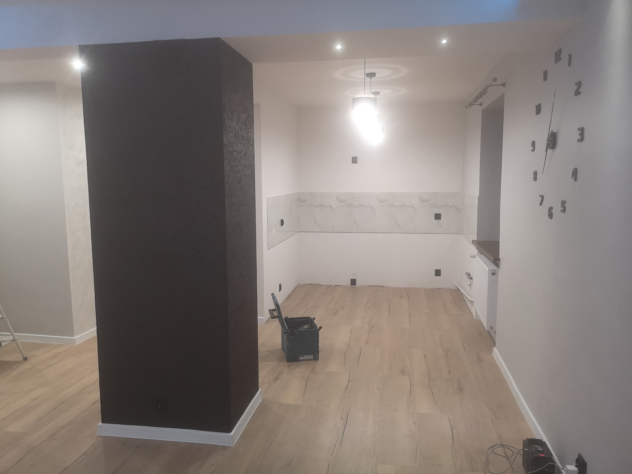 Wnętrze w trakcie prac wykończeniowych: drewnopodobna podłoga, pomalowany filar na ciemny brąz, częściowo wykończona ściana z płytkami imitującymi marmur.