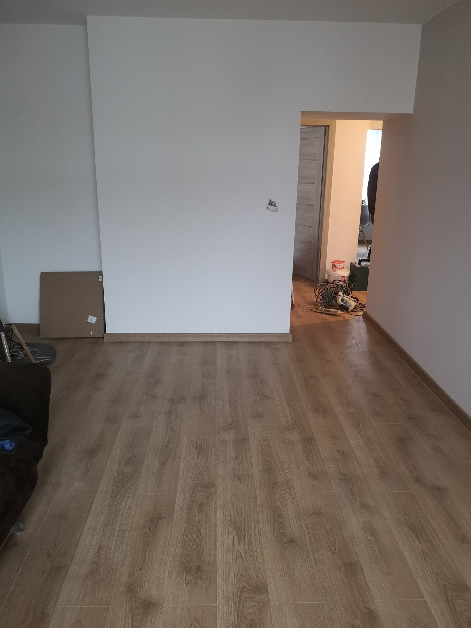 Jasne panele podłogowe w pokoju po remoncie, widoczne narzędzia i materiały w przejściu do kolejnego pomieszczenia. Ściany pomalowane na biało.