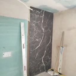 Montaż płytek imitujących marmur na ścianie w łazience w trakcie remontu. Widoczne narzędzia, poziomica i płyta karton gips na ścianach.