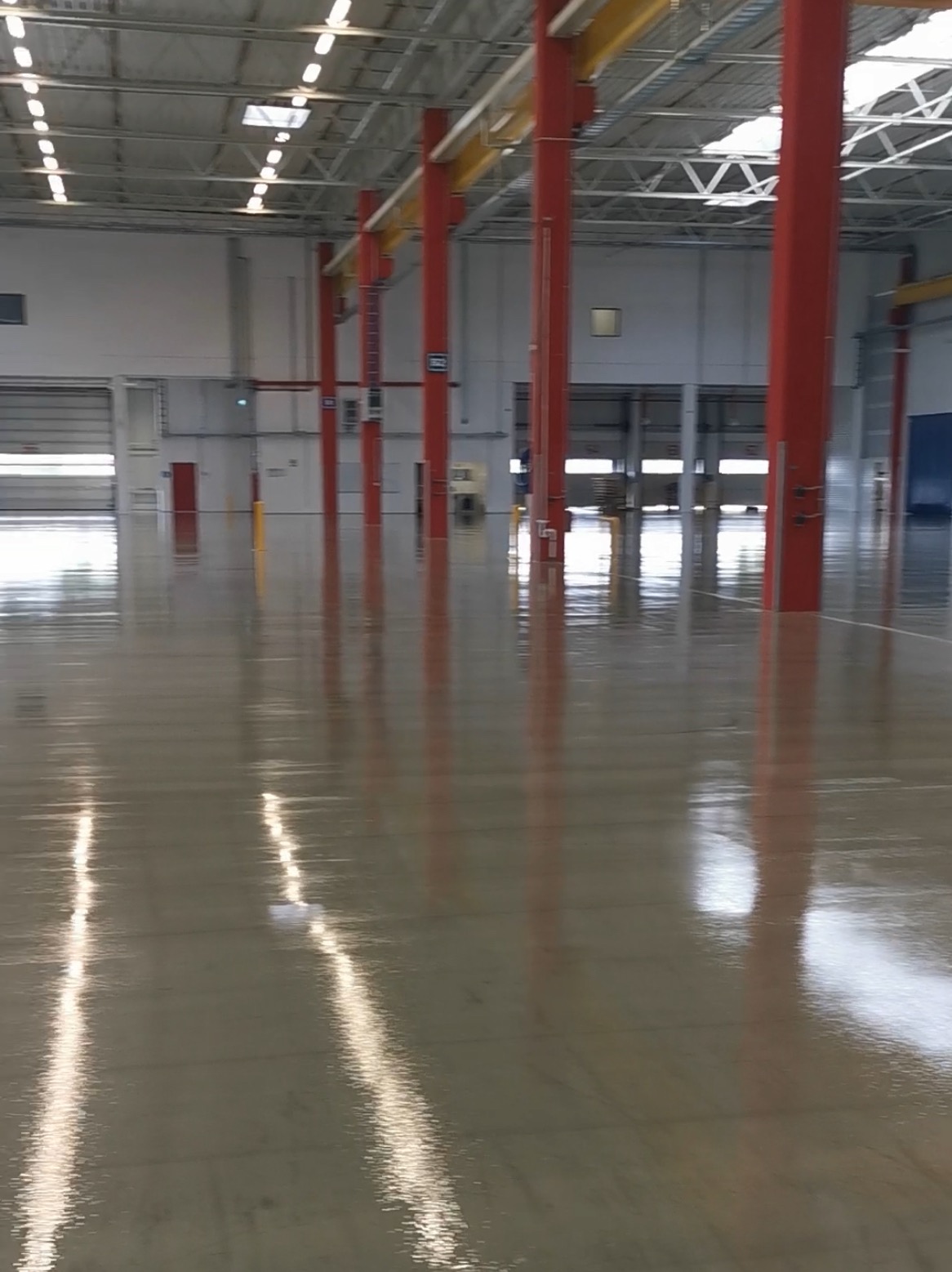 Błyszcząca, beżowa posadzka w hali przemysłowej z czerwonymi kolumnami. Widoczne odbicia światła w gładkiej powierzchni, podkreślające jej równość i estetykę.