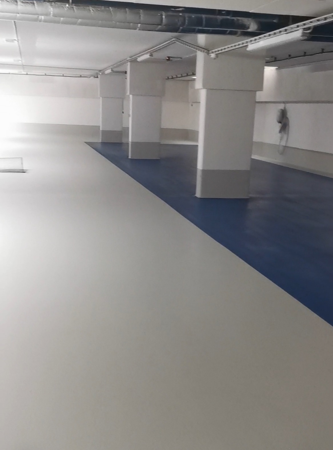 Jasna, dwukolorowa posadzka w hali z kolumnami. Część powierzchni w kolorze białym, część w kolorze niebieskim. Widoczne oświetlenie i instalacje pod sufitem.