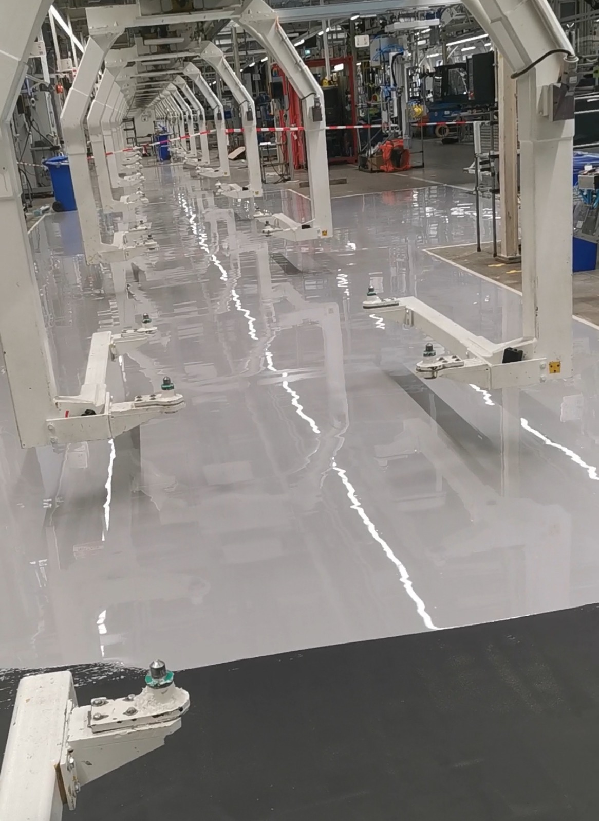 Błyszcząca, szara posadzka w hali produkcyjnej z rzędami białych maszyn. Gładka powierzchnia odbija światło lamp, tworząc efekt lustrzany. Widok z perspektywy podłogi.