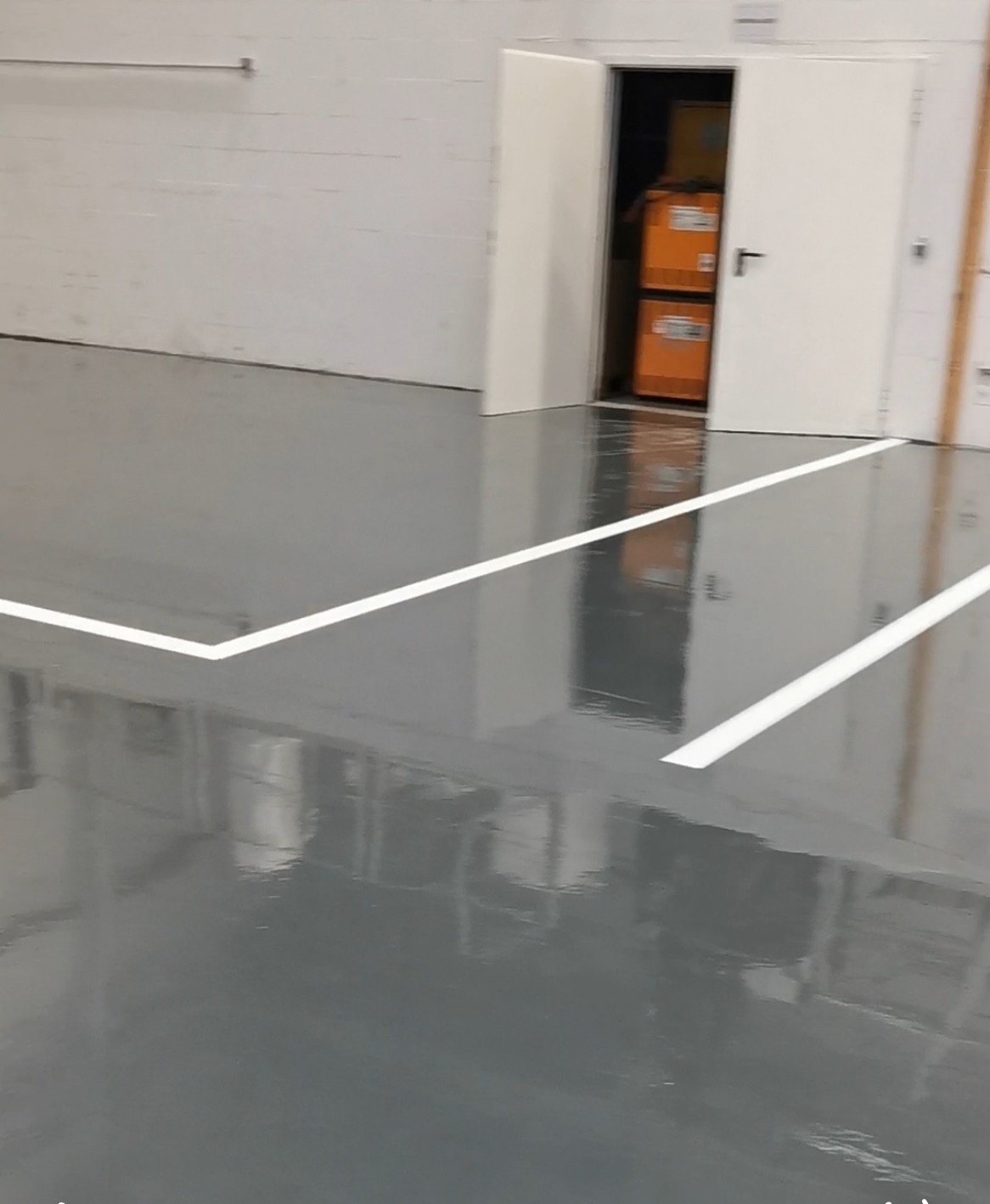 Szara, błyszcząca posadzka przemysłowa z białymi liniami wyznaczającymi strefy, widok na otwarte drzwi z magazynowanymi przedmiotami w tle.